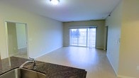 This single-floor space showcases a clean, open-concept layout that seamlessly connects the kitchen and living areas. The living room is finished with light hardwood-style flooring and neutral-toned walls, creating a bright and versatile environment. A large sliding glass door dominates the far wall, allowing generous natural light to fill the space while providing direct access to a balcony with an outdoor view of surrounding greenery. The balcony features a railing system, making it a functional extension of the interior living area.

The kitchen, partially visible in the foreground, features rich, dark granite countertops that contrast elegantly with the lighter tones throughout the rest of the space. A sink with a contemporary faucet is integrated into the countertop, adding a practical yet polished touch.

Ceiling-mounted lighting provides even illumination throughout the living area, and multiple electrical outlets are positioned along the baseboards for convenience. The overall design aesthetic is modern and understated, with an emphasis on open space, natural light, and clean finishes that offer a comfortable and functional living environment.