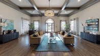 This elegantly appointed single-floor community space exudes a refined, welcoming ambiance from the moment you enter. The grand focal point of the main lounge is a stunning arched double-door entryway flanked by tall windows dressed in bold patterned drapes, flooding the room with natural light. Overhead, a striking coffered ceiling with dark-stained beams adds architectural depth and sophistication, complemented by a statement drum chandelier that anchors the seating area below. Rich hardwood-style flooring runs throughout, tying the space together seamlessly. The primary lounge features two generously sized tufted sofas in warm neutral tones, accented with teal and gold throw pillows, arranged around a glass-top coffee table atop a vivid teal area rug. Lush potted plants add natural warmth to the corners. A secondary seating area showcases a dramatic floor-to-ceiling fireplace with a sleek white surround adorned with decorative metallic horizontal banding, paired with tall sculptural vases and a wingback accent chair. Additional areas include a casual lounge with open sightlines to adjacent rooms, a sideboard with refreshment amenities, and curated artwork on the walls. The overall design balances contemporary elegance with inviting comfort.