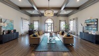 This elegantly appointed single-floor community space exudes a refined, welcoming ambiance from the moment you enter. The grand focal point of the main lounge is a stunning arched double-door entryway flanked by tall windows dressed in bold patterned drapes, flooding the room with natural light. Overhead, a striking coffered ceiling with dark-stained beams adds architectural depth and sophistication, complemented by a statement drum chandelier that anchors the seating area below. Rich hardwood-style flooring runs throughout, tying the space together seamlessly. The primary lounge features two generously sized tufted sofas in warm neutral tones, accented with teal and gold throw pillows, arranged around a glass-top coffee table atop a vivid teal area rug. Lush potted plants add natural warmth to the corners. A secondary seating area showcases a dramatic floor-to-ceiling fireplace with a sleek white surround adorned with decorative metallic horizontal banding, paired with tall sculptural vases and a wingback accent chair. Additional areas include a casual lounge with open sightlines to adjacent rooms, a sideboard with refreshment amenities, and curated artwork on the walls. The overall design balances contemporary elegance with inviting comfort.