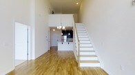 This two-story space makes a strong first impression with its dramatic double-height ceilings that flood the main living area with a sense of openness and volume. Light hardwood flooring runs continuously throughout the main level, lending warmth and cohesion to the open-concept layout.

A elegant staircase with white-painted risers and a natural wood handrail anchors the space, rising gracefully to the upper level while serving as a visual focal point. The generous ceiling height above allows the main living area to feel expansive and airy.

Toward the back of the main level, the kitchen is defined by dark cabinetry that creates a striking contrast against the neutral walls and light flooring. A white kitchen island with a breakfast bar provides both a functional workspace and casual dining option. Stainless steel appliances complete the kitchen's contemporary look, while pendant lighting adds a refined finishing touch overhead.

A chandelier hangs centrally in the main living area, complementing the recessed lighting above to ensure the space is well-lit throughout. The upper level overlooks the main floor, reinforcing the home's open, connected feel.