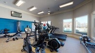 This fitness center is a comprehensively outfitted facility designed to support a full range of workout routines. The space is finished with neutral gray walls accented by a bold blue feature wall, crown molding, and durable low-pile carpet flooring throughout, creating a clean and energizing environment.

The cardio zone is well-stocked with multiple treadmills, elliptical cross-trainers, a stationary bike, and a rowing machine, all arranged to maximize usability. A ceiling fan and flush-mounted lighting fixtures keep the space comfortable and well-lit. Large windows along one wall bathe the room in natural light while offering views of an outdoor pool area, adding a refreshing visual element during workouts.

The strength training area is equally impressive, featuring a multi-station cable machine, a weight bench with press attachments, a lat pulldown tower, and additional resistance machines. A rack of medicine balls and stability balls provides options for functional and core training. Floor-to-ceiling mirrors line one section of the space, visually expanding the room and allowing users to monitor their form during exercises. Wall-mounted flat-screen televisions are positioned for easy viewing throughout the facility. A door leads directly to the outdoor pool area, offering convenient access between indoor and outdoor amenities.