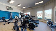 This fitness center is a comprehensively outfitted facility designed to support a full range of workout routines. The space is finished with neutral gray walls accented by a bold blue feature wall, crown molding, and durable low-pile carpet flooring throughout, creating a clean and energizing environment.

The cardio zone is well-stocked with multiple treadmills, elliptical cross-trainers, a stationary bike, and a rowing machine, all arranged to maximize usability. A ceiling fan and flush-mounted lighting fixtures keep the space comfortable and well-lit. Large windows along one wall bathe the room in natural light while offering views of an outdoor pool area, adding a refreshing visual element during workouts.

The strength training area is equally impressive, featuring a multi-station cable machine, a weight bench with press attachments, a lat pulldown tower, and additional resistance machines. A rack of medicine balls and stability balls provides options for functional and core training. Floor-to-ceiling mirrors line one section of the space, visually expanding the room and allowing users to monitor their form during exercises. Wall-mounted flat-screen televisions are positioned for easy viewing throughout the facility. A door leads directly to the outdoor pool area, offering convenient access between indoor and outdoor amenities.