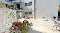 This ground-floor unit is situated within a multi-story residential building characterized by a clean, modern exterior finished in crisp white stucco with dark-framed windows. The building's courtyard-style layout creates a semi-private outdoor environment shared among residents, featuring a spacious concrete patio that receives generous natural sunlight throughout the day.

The courtyard is well-appointed with an array of potted trees, flowering shrubs, and layered plantings that add color and texture to the otherwise minimalist architectural palette. Decorative white lattice panels serve as privacy dividers within the courtyard, creating distinct zones while maintaining an open, airy feel. Seating areas with lounge chairs and small accent tables are positioned throughout, encouraging outdoor relaxation.

A separate exterior patio area, accessible via iron railing fencing, offers additional outdoor seating with stone-topped tables and views toward neighboring greenery and a mature palm tree. A barbecue grill is also present, adding functional outdoor living appeal.

Upper-level balconies overlook the courtyard, contributing to a layered, community-oriented atmosphere. Climbing vines, papyrus, and flowering plants add organic detail against the smooth white walls. The overall ambiance is bright, tranquil, and well-maintained, with a strong emphasis on outdoor living and greenery.