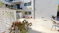 This ground-floor unit is situated within a multi-story residential building characterized by a clean, modern exterior finished in crisp white stucco with dark-framed windows. The building's courtyard-style layout creates a semi-private outdoor environment shared among residents, featuring a spacious concrete patio that receives generous natural sunlight throughout the day.

The courtyard is well-appointed with an array of potted trees, flowering shrubs, and layered plantings that add color and texture to the otherwise minimalist architectural palette. Decorative white lattice panels serve as privacy dividers within the courtyard, creating distinct zones while maintaining an open, airy feel. Seating areas with lounge chairs and small accent tables are positioned throughout, encouraging outdoor relaxation.

A separate exterior patio area, accessible via iron railing fencing, offers additional outdoor seating with stone-topped tables and views toward neighboring greenery and a mature palm tree. A barbecue grill is also present, adding functional outdoor living appeal.

Upper-level balconies overlook the courtyard, contributing to a layered, community-oriented atmosphere. Climbing vines, papyrus, and flowering plants add organic detail against the smooth white walls. The overall ambiance is bright, tranquil, and well-maintained, with a strong emphasis on outdoor living and greenery.