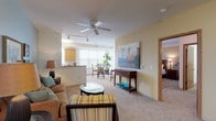 This single-floor residence offers a thoughtfully designed layout that balances comfort and functionality. The open-concept living and dining area serves as the heart of the home, featuring soft carpeting, warm neutral wall tones, and a ceiling fan with track lighting. Large windows dressed with vertical blinds flood the space with natural light, creating an airy and welcoming atmosphere. A glass-top dining table with seating for four anchors the dining area, which flows seamlessly into the living room, accented by a teal feature wall and curated wall art.

The galley-style kitchen is well-equipped with ample honey-toned cabinetry, black appliances including a refrigerator, range, microwave, and dishwasher, along with generous counter space and a double sink beneath a window. Wood-look flooring adds warmth and practicality to this functional space.

The primary bedroom is generously sized with plush carpeting, a ceiling fan, and warm lighting, complemented by an en suite bathroom featuring a wood-vanity, white countertop, and a bathtub. The secondary bedroom offers carpeted floors and a built-in closet with sliding oak doors. A second full bathroom with wood-look flooring and a single vanity serves this room. A welcoming entry foyer with an oak panel door completes the layout.