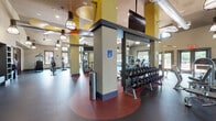 This fully equipped fitness center at 500 Bristol Blvd in Somerset, NJ, offers a comprehensive workout environment spread across a single floor. The space is architecturally dynamic, featuring soaring vaulted ceilings, exposed ductwork, and bold yellow, red, and orange ceiling accents that give the facility a modern, energetic character. Pendant lighting throughout complements abundant natural light streaming in through large arched and rectangular windows, providing views of the surrounding greenery.

The cardio zone is generously appointed with multiple treadmills, elliptical machines, and stationary bikes, all positioned to take advantage of the natural light and outdoor views. A dedicated row of spinning bikes occupies its own area, complete with a flat-screen TV, framed artwork, and built-in shelving. The free weights section features a full dumbbell rack set on a distinctive red rubberized floor, accompanied by adjustable benches and a wall of mirrors. The strength and selectorized machine zone includes a variety of cable and resistance machines arranged across a spacious, dark rubberized floor. Ceiling-mounted televisions provide entertainment throughout, and the facility's open layout ensures ample room for movement and flexibility training.