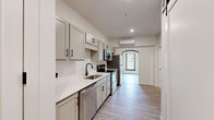 This thoughtfully renovated single-floor unit offers a clean, contemporary aesthetic across a well-organized layout. The galley-style kitchen serves as a central design feature, showcasing sage-gray shaker cabinets with matte black hardware, white quartz countertops, a single-basin sink with a black gooseneck faucet, and a full suite of stainless steel appliances including a refrigerator, electric range, built-in microwave, and dishwasher. A wall-mounted mini-split HVAC unit ensures year-round climate control. An arched passage leads from the kitchen into the open living area, which is bathed in natural light from two large, dark-trimmed windows overlooking the surrounding neighborhood. The living space features crisp white walls, light wood-look vinyl plank flooring, and an accent of exposed brick visible near the entryway, adding character to the otherwise modern interior. The bathroom is finished with a white vanity, matching quartz countertop, and a walk-in shower with white subway-style tile and matte black fixtures. A dedicated in-unit laundry closet with a front-loading washer adds practical convenience. A sliding barn door and a bold yellow entry door contribute distinctive design touches throughout.