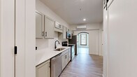 This thoughtfully renovated single-floor unit offers a clean, contemporary aesthetic across a well-organized layout. The galley-style kitchen serves as a central design feature, showcasing sage-gray shaker cabinets with matte black hardware, white quartz countertops, a single-basin sink with a black gooseneck faucet, and a full suite of stainless steel appliances including a refrigerator, electric range, built-in microwave, and dishwasher. A wall-mounted mini-split HVAC unit ensures year-round climate control. An arched passage leads from the kitchen into the open living area, which is bathed in natural light from two large, dark-trimmed windows overlooking the surrounding neighborhood. The living space features crisp white walls, light wood-look vinyl plank flooring, and an accent of exposed brick visible near the entryway, adding character to the otherwise modern interior. The bathroom is finished with a white vanity, matching quartz countertop, and a walk-in shower with white subway-style tile and matte black fixtures. A dedicated in-unit laundry closet with a front-loading washer adds practical convenience. A sliding barn door and a bold yellow entry door contribute distinctive design touches throughout.