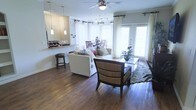 This single-story residence features an open-concept layout that connects the living, dining, and kitchen areas into one cohesive and functional space. Rich, dark hardwood flooring runs throughout, adding warmth and a sense of continuity across the open floor plan.

The living area is comfortably sized and anchored by a ceiling fan with integrated lighting. Large windows dressed with white curtains and horizontal blinds allow generous natural light to fill the room, while a set of glass French doors provides access to the outdoors and further brightens the space.

A woven accent chair and a plush white sofa with decorative pillows are arranged around a glass-top coffee table set atop a textured green area rug, creating an inviting seating arrangement. Adjacent to the living area, a round dining table with wood chairs occupies a windowed nook, offering a pleasant spot for everyday meals.

The kitchen is partially visible beyond a raised breakfast bar with pendant lighting overhead, offering a casual seating option and a natural visual divide between the kitchen and living areas. The overall ambiance is light, airy, and welcoming.