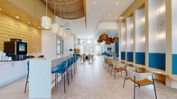 This single-floor amenity clubhouse, branded "Perla," offers a generously proportioned, open-plan layout that seamlessly blends social, lounge, and café functions into one cohesive environment. The interior design is defined by a coastal-contemporary aesthetic, featuring a warm palette of sandy neutrals, bold teal and cobalt blue accents, and rich golden tones.

A central café bar with a striking geometric-tiled base and teal cabinetry serves as a focal point, illuminated by an overhead cluster of oversized woven rattan pendant lights. Bar-height seating with teal upholstered chairs lines a long white counter, creating a lively social hub. Adjacent open shelving and a neon "Perla" sign add a distinctive, curated character.

The lounge area features a thoughtful mix of seating styles, from sculptural low tables with modern chairs to a blue sofa adorned with teal cushions, accented by starburst wall sconces. A dramatic architectural wall of arched, illuminated niches with teal lower panels provides a stunning backdrop to casual seating arrangements nearby.

Near the entrance, a double-height foyer showcases circular pod seating built into a slatted wood accent wall, with floor-to-ceiling glass doors opening to outdoor pool views. Recessed lighting and large windows fill the entire space with natural light. Access to a dedicated fitness center completes this well-appointed amenity suite.