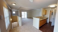 This single-floor space showcases an open-concept layout that seamlessly connects the kitchen and living areas. The vaulted ceilings with crown molding add a sense of height and architectural interest, while flush-mount lighting provides functional illumination throughout.

The kitchen features warm honey-toned cabinetry paired with granite countertops and an integrated sink, positioned along a peninsula that serves as a natural divider between the kitchen and living spaces. The flooring transitions from dark hardwood in the kitchen to plush carpeting in the living area, creating a clear visual distinction between the two zones.

The living area is bright and open, benefiting from natural light provided by a sliding glass door that opens onto an outdoor balcony or deck, as well as an additional window on the adjacent wall. The neutral wall tones and light carpeting contribute to an airy, welcoming ambiance.

A staircase is visible at the edge of the space, suggesting access to lower levels. Crown molding throughout ties the design together, lending a polished, finished look to this well-appointed, light-filled space.