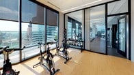 This contemporary fitness center is thoughtfully designed to support a comprehensive range of workout routines across a single, open floor. The main exercise area is lined with floor-to-ceiling windows that flood the space with natural light and offer sweeping city and skyline views, creating an energizing atmosphere for training. The flooring throughout the primary zone features durable rubber-speckled material, ideal for high-traffic fitness use.

The cardio section is well-appointed with a row of treadmills, elliptical machines, stair climbers, and stationary bikes, all positioned to take advantage of the panoramic exterior views. Adjacent to the cardio area, a dedicated strength training zone houses a variety of cable machines, weight benches, and selectorized equipment, complemented by wall-mounted mirrors and flat-screen TVs.

A separate enclosed studio provides space for spin cycling, with multiple indoor bikes and an array of kettlebells and medicine balls for functional training. Plyosoft boxes and step platforms make this studio suitable for high-intensity interval and agility workouts. The studio features light wood flooring and large windows, adding warmth and brightness. A dedicated corner area includes open shelving, artwork, and additional strength machines, giving the space an organized, boutique-gym feel throughout.