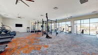 Located at 3875 W Dobbins Rd in Phoenix, AZ, this professionally designed fitness center offers a comprehensive, single-floor exercise facility that covers a generously sized footprint. The main workout area is anchored by a multi-station functional rig equipped with TRX suspension trainers, battle ropes, and a heavy bag, set atop a striking orange and grey patterned carpet. Large floor-to-ceiling windows line the perimeter, flooding the space with natural light and providing views of the surrounding palm-lined outdoor area. Ceiling fans are positioned throughout to maintain airflow and comfort. The cardio zone features treadmills, ellipticals, and air bikes arranged along the windowed wall, while a dedicated free weight and strength section includes power racks, adjustable benches, and a full dumbbell rack backed by floor-to-ceiling mirrors. A separate studio room showcases an elegant herringbone-patterned hardwood floor, a calming watercolor mural accent wall, and a built-in display screen set against a teal accent wall — ideal for group fitness, yoga, or personal training sessions. A tiled entry zone with a weight bench and power rack provides a transitional strength training area near the exterior access door, completing this well-appointed, versatile fitness facility.