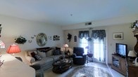 This single-floor space offers a cozy and classically styled living environment thoughtfully curated with traditional décor. The living area serves as the heart of the home, centered around a cushioned sofa and matching armchair upholstered in neutral tones, complemented by an elegant oval coffee table that anchors the space. A plush area rug with floral detailing adds warmth and texture underfoot, while light-toned flooring extends throughout.

Decorative touches abound, including a vintage-style round mirror adorned with floral motifs mounted on the wall, delicate butterfly wall accents, and a Tiffany-style table lamp that casts a soft ambient glow. Wall shelving displays curated collectibles, adding a personalized charm to the room.

Natural light flows generously into the space through a large glass door dressed with layered fabric valances in soft, muted tones, offering a view to the outside. Venetian blinds provide flexible light control. A wall-mounted HVAC vent and a ceiling-mounted sprinkler point to practical, well-maintained systems throughout. The smooth white ceilings and neutral walls create a clean backdrop that lets the furnishings and décor take center stage, resulting in an inviting and comfortable atmosphere.