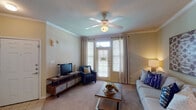 This single-floor 1-bedroom, 1-bathroom residence offers a thoughtfully designed layout that maximizes comfort and functionality. The living room serves as the heart of the home, featuring crown molding, warm neutral-toned walls, and plush carpeting. A ceiling fan overhead provides year-round comfort, while a French door flanked by large windows with elegant drapery fills the space with natural light and provides outdoor access. The open feel is enhanced by a smooth transition from tile at the entry to carpet throughout the main living areas. The galley-style kitchen is equipped with warm wood cabinetry, a full suite of white appliances including a range, over-the-range microwave, and refrigerator, complemented by tile flooring and laminate countertops. A dedicated dining area connects seamlessly to the kitchen and living space. The bedroom is a serene retreat, highlighted by a ceiling fan, a generous window dressed with bright curtains, and soft carpet underfoot. The full bathroom features a tub-shower combination, a rich wood vanity with a large mirror, and tile flooring. Crown molding and consistent neutral tones tie the entire residence together with a cohesive, welcoming aesthetic.