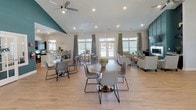 This single-floor clubhouse is designed to serve as a welcoming community hub, characterized by soaring vaulted ceilings and a bold teal accent wall that anchors the open interior. Light hardwood flooring runs throughout, contributing to a bright and cohesive aesthetic. Recessed lighting and a ceiling fan with light fixture provide consistent illumination across the main gathering area.

The social lounge area features a comfortable sectional sofa, a yellow upholstered ottoman, and coordinating accent furniture arranged atop a patterned area rug, creating an inviting space for relaxed conversation. Adjacent to this is a dining and café-style seating area furnished with round tables and modern grey chairs, ideal for casual meetings or events.

A dedicated leasing office is separated from the main floor by elegant French doors with glass panes, providing a semi-private workspace with a desk, seating, and ample natural light from large windows dressed in patterned curtains. The office connects seamlessly to the main gathering space, maintaining an open and accessible feel.

Multiple windows along the perimeter flood the interior with natural light and offer views of the surrounding greenery, enhancing the overall airy ambiance of the clubhouse.