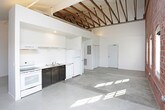 This single-floor loft-style space showcases an impressive blend of industrial character and modern functionality. The main living area is defined by striking exposed red brick walls, soaring vaulted ceilings with visible timber roof trusses, and polished concrete floors that reflect natural light throughout. A row of arched windows lines one wall, flooding the open space with abundant daylight and offering views of the surrounding greenery, enhancing the airy, expansive atmosphere.

The compact kitchen is thoughtfully integrated into the open floor plan, featuring a white range, refrigerator, and dark cabinetry beneath a simple open shelf. A sliding door leads to additional storage or utility space nearby, while the exposed structural elements overhead lend a raw, loft-inspired aesthetic.

The bathroom is clean and efficiently designed, outfitted with a full bathtub and shower combination, a toilet, and a dark-framed vanity with a mirror. A practical stacked Whirlpool washer and dryer unit is conveniently housed within the bathroom, maximizing the use of space. The consistent polished concrete flooring carries throughout, tying the space together with a cohesive, industrial-modern feel. Overall, the space offers a distinctive living environment rich in architectural character.