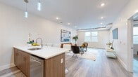 This thoughtfully designed one-bedroom, one-bathroom residence offers a bright and modern open-concept layout with clean lines and contemporary finishes throughout. Light grey wood-style flooring flows seamlessly across the entire unit, anchoring the cohesive aesthetic.

The kitchen is a standout feature, showcasing walnut-toned cabinetry, crisp white quartz countertops, full-suite stainless steel appliances, and a center island with bar seating for three — illuminated by sleek cylindrical pendant lights. The kitchen opens directly into the dining and living area, creating an expansive, sociable space bathed in natural light from large windows at the far end.

The living room is generously proportioned, offering ample room for comfortable seating arrangements, and is accented by recessed lighting and a neutral color palette that enhances the airy atmosphere.

The bedroom is a serene retreat, featuring a walk-in closet, a natural light source from its own window, and enough space to accommodate a king-sized bed with ease. Subtle exposed ductwork adds a modern industrial touch to the room.

The bathroom features a full soaking tub framed by striking marble-patterned wall surrounds with elegant grey veining and polished chrome fixtures, lending a spa-like quality to the space.