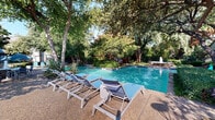 Located at 9821 Summerwood Cir in Dallas, Texas, this outdoor amenity space offers a resort-inspired pool experience nestled within a beautifully landscaped environment. The centerpiece of the area is a generously sized, sparkling swimming pool with clear blue water, complemented by a separate wading or fountain pool that adds a dynamic visual element to the setting.

The pool deck is finished with a textured concrete surface and features tiered stone retaining walls that give the area a sense of depth and dimension. Mature oak and crape myrtle trees — including a striking specimen with vibrant pink blooms — provide generous natural shade throughout the space, creating a cool and comfortable outdoor retreat even on warm Texas days.

Scattered across the deck are multiple lounge chairs and seating arrangements, including umbrella-shaded tables ideal for outdoor dining or socializing. A pergola structure is positioned along the perimeter, offering additional shaded seating and lending a refined architectural touch to the grounds. Lush native plantings, agaves, and manicured shrubs frame the pool area, enhancing its natural ambiance. An adjacent building with large glass doors provides convenient access to interior facilities. The overall atmosphere is tranquil, verdant, and well-appointed for outdoor leisure.