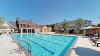 This single-level outdoor amenity space is centered around a generously sized swimming pool with clearly marked lap lanes and crystal-clear blue water, ideal for both recreational swimming and fitness. The pool deck is finished with clean, light-toned pavers and is equipped with multiple entry points via stainless steel handrails and ladders. Surrounding the pool area, a classic brick and neutral-toned clubhouse structure provides a backdrop, complemented by large windows and covered patio sections with orange-accented umbrellas and outdoor seating arrangements.

Adjacent to the pool, elegantly designed cabanas offer a private retreat from the sun. Each cabana features a canopied roof, flowing neutral drape curtains that can be drawn for privacy, and plush wicker lounge seating accented with vibrant orange and cream cushions. The cabanas overlook carefully maintained garden beds filled with colorful ornamental plantings, including deep red and green foliage that adds a lush, vibrant character to the surroundings.

The entire amenity area is enclosed by a sleek wrought iron fence, providing both security and an open sightline to the surrounding residential buildings. The overall ambiance is polished, inviting, and resort-inspired, offering a high-quality outdoor living experience.