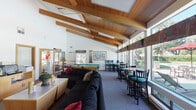 Located at 1420 F St in Davis, California, this single-story Clubhouse and Fitness Center serves as a well-appointed community gathering space designed for both relaxation and activity. The main lounge area is the centerpiece of the facility, boasting soaring vaulted ceilings with warm exposed wood beams and a striking wood-paneled ceiling that lends a welcoming, lodge-like ambiance. Large windows along one wall flood the space with natural light and offer views of the surrounding greenery. The lounge is comfortably furnished with ample seating, including a generous sectional sofa, and features a decorative tiled fireplace with a colorful teal accent wall — a distinctive architectural element that adds character to the room. A pool table provides a focal point for recreation, complemented by high-top bistro tables and bar-height seating arranged along the windowed wall. The kitchenette area offers practical amenities including upper and lower cabinetry, a microwave, coffee maker, and sink — ideal for community events. The dedicated fitness room is equipped with multiple cardio machines, including treadmills and ellipticals, and benefits from its own natural light and wood-beamed ceiling. Additional strength and exercise equipment rounds out the workout offerings, making this a comprehensive and inviting community amenity.