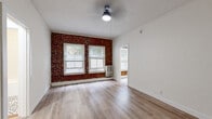 This single-floor unit blends classic character with modern updates across a thoughtfully arranged layout comprising a living room, kitchen, bathroom, and hallway. The living room serves as the centerpiece of the space, showcasing a striking exposed red brick accent wall with arched window details that add historic charm. Two windows with blinds invite natural light, while light-toned hardwood flooring and a ceiling fan overhead contribute to a clean, airy atmosphere. The opposite side of the living area features crisp white walls and an entry door, maintaining a bright and open feel throughout. The kitchen is efficiently designed with white shaker-style cabinetry, light stone countertops, a stainless steel single-basin sink, and a stainless steel gas range with an overhead ventilation hood — offering both style and function in a compact footprint. The bathroom stands out with its classic black-and-white patterned tile flooring, white subway tile surround in the tub-shower combination, a pedestal sink, and a recessed medicine cabinet mirror — delivering a timeless, well-appointed finish. The hallway connects the rooms seamlessly, maintaining the consistent hardwood flooring found throughout. Overall, the space presents a cohesive blend of vintage architectural details and contemporary finishes.