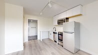 This single-floor unit offers a clean, modern design centered around an open-concept layout that maximizes functionality and flow. Light hardwood flooring runs throughout the main living area, complemented by crisp white walls and track lighting that illuminate the space with a bright, airy ambiance.

The kitchen is well-appointed with white shaker-style cabinetry, a stainless steel range, built-in microwave, dishwasher, and a full-size refrigerator. A compact countertop peninsula adds prep space and defines the kitchen zone within the open floor plan.

The main living area is spacious and versatile, featuring an accessible electrical panel recessed into the wall and a dedicated built-in closet with two shelving tiers, providing practical storage. A doorway leads directly to the bathroom, creating a convenient layout.

The bathroom is finished with large-format dark stone-look floor tiles, offering a striking contrast to the white walls. It includes a single-sink vanity with a modern matte black faucet, a round mirror with black framing, and matching black hardware accents throughout, lending a contemporary, cohesive aesthetic.

Overall, this unit presents a thoughtfully designed space that balances minimalist style with everyday practicality, ideal for those seeking a modern, move-in-ready environment.