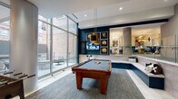This contemporary amenity space is thoughtfully designed across a single level, offering a dynamic mix of recreational and social areas that cater to a variety of activities. The game room serves as a focal point, anchored by a full-size billiards table set atop a textured area rug, complemented by a foosball table nearby. A dramatic navy blue accent wall adorned with framed artwork and a wall-mounted television adds bold character to the space. Adjacent to the game area, a built-in L-shaped banquette with cushioned seating and decorative pillows provides a casual gathering spot, partially separated from a neighboring lounge by a glass-railed half-wall. Floor-to-ceiling windows along one wall flood the interior with abundant natural light while offering views of an adjacent outdoor terrace. A modern fireplace with a sleek surround anchors a comfortable seating arrangement, while pendant lighting in warm metallic tones adds an elegant touch throughout. The lounge area features plush, curved sofas and eclectic accent furniture beneath a gold pendant light, separated from a communal workspace by a full-height glass partition. The lobby entry area is finished with large-format light stone tile flooring, a reception counter, pendant lights, and a beverage station with a patterned tile backsplash, completing this well-rounded and inviting amenity level.