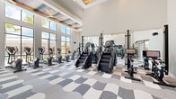 Located at 2525 Paseo Park Rd in Orlando, Florida, this contemporary fitness center is a well-appointed, single-floor facility designed to support a full range of workout routines. The space is defined by its soaring double-height ceilings, which are accented by coffered details and large ceiling fans that keep the environment comfortable throughout. Floor-to-ceiling windows line the perimeter, flooding the interior with abundant natural light and offering sweeping views of the surrounding palm-lined landscape. The flooring throughout features a bold, wavy geometric carpet pattern in shades of gray and white, adding a dynamic visual character to the space. The main workout area is generously sized and organized into distinct zones. A dedicated cardio section houses a lineup of treadmills, elliptical trainers, stair climbers, and stationary bikes, all positioned to take advantage of the outdoor views. The strength training zone is equipped with power racks, weight benches, free weights, and plate-loaded machines. Full-length mirrors are strategically placed along the walls to enhance the sense of space and support proper form. A glass-enclosed room provides a separate training area with turf flooring, ideal for functional fitness and agility work. The overall ambiance is bright, modern, and energizing.