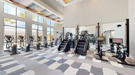 Located at 2525 Paseo Park Rd in Orlando, Florida, this contemporary fitness center is a well-appointed, single-floor facility designed to support a full range of workout routines. The space is defined by its soaring double-height ceilings, which are accented by coffered details and large ceiling fans that keep the environment comfortable throughout. Floor-to-ceiling windows line the perimeter, flooding the interior with abundant natural light and offering sweeping views of the surrounding palm-lined landscape. The flooring throughout features a bold, wavy geometric carpet pattern in shades of gray and white, adding a dynamic visual character to the space. The main workout area is generously sized and organized into distinct zones. A dedicated cardio section houses a lineup of treadmills, elliptical trainers, stair climbers, and stationary bikes, all positioned to take advantage of the outdoor views. The strength training zone is equipped with power racks, weight benches, free weights, and plate-loaded machines. Full-length mirrors are strategically placed along the walls to enhance the sense of space and support proper form. A glass-enclosed room provides a separate training area with turf flooring, ideal for functional fitness and agility work. The overall ambiance is bright, modern, and energizing.