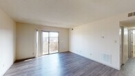 This single-floor unit offers a practical and well-organized layout across its main living spaces. The living room serves as the centerpiece of the unit, featuring smooth hardwood-style flooring, neutral cream walls, and a textured ceiling. A large sliding glass door floods the space with natural light and provides direct access to a private balcony with outdoor views, enhancing the open and airy feel of the room. Adjacent to the living area, the kitchen is equipped with dark wood cabinetry, black appliances including a refrigerator and range, and tile flooring that transitions neatly into the main living space. Overhead lighting illuminates the kitchen efficiently. A hallway connects the living area to the private quarters of the unit. The bedroom is generously sized, featuring plush carpeting, a large window dressed with vertical blinds that allow ample natural light, and a louvered double-door closet offering practical storage. The bathroom is well-appointed with an extended countertop, dark wood cabinetry beneath the vanity, a well-lit mirror above the sink, tile flooring, and a separate shower area. Throughout the unit, the consistent use of neutral tones and functional finishes creates a clean, understated ambiance.