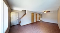 This two-story space presents a practical and open main level anchored by warm, dark hardwood-style flooring that extends throughout the area. A staircase with a wooden handrail leads to the upper floor and is positioned adjacent to the front entry door, which is finished in classic white. The main level features neutral gray walls and a textured ceiling, contributing to a clean and understated aesthetic. A ceiling fan is installed overhead, providing both lighting and ventilation to the living area. Along the main wall, two medium-toned wooden doors provide access to additional spaces, while a hallway toward the rear of the unit leads further into the home, where a white appliance — likely a refrigerator — is partially visible, indicating the kitchen or utility area. A wall-mounted heating unit is also visible along the baseboard, suggesting built-in climate control. The layout flows openly across the main floor, offering a versatile living space. Overall, the design is straightforward and functional, with warm wood tones providing contrast against the light walls and ceiling throughout.