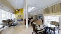This single-story clubhouse offers an expansive, open-concept layout designed to foster community and relaxation. The interior is immediately striking thanks to its soaring vaulted ceilings, which are enhanced by skylights that flood the space with natural light throughout the day. Ceiling fans are strategically placed to ensure comfortable airflow across the generous floor plan.

The main gathering area is anchored by multiple seating arrangements, including plush sofas and lounge chairs grouped around a central media area with a large flat-screen television. A bold yellow accent wall adds a vibrant, energetic contrast to the neutral beige and cream tones that dominate the space. Smooth tile flooring runs throughout, lending a clean, cohesive aesthetic that is both practical and elegant.

Structural columns help define distinct zones within the open floor plan without compromising the sense of spaciousness. A dining area with a glass-top table and woven chairs provides a comfortable spot for casual meals or conversation. Large windows dressed with shutters and striped Roman shades allow for adjustable natural light while maintaining privacy. The overall ambiance is warm, inviting, and well-appointed, making this clubhouse an ideal venue for community gatherings and everyday leisure.