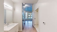 Located at 1741 36th St in Denver, Colorado, this single-floor unit presents a clean, contemporary design characterized by a bright, neutral palette and light wood-style flooring throughout. The open layout connects functional living areas efficiently, creating a cohesive flow from entry to living space.

Upon entry, a vanity with a white countertop and large mirror is immediately visible alongside the front door, which features a distinctive natural wood finish with a navy blue frame. A closet sits nearby, adding practical storage at the threshold.

The kitchen is compact yet well-equipped, outfitted with stainless steel appliances including a refrigerator, electric range, over-the-range microwave, and dishwasher. White cabinetry and open shelving maximize storage, while a stainless steel sink and white countertops maintain the modern aesthetic.

The main living area benefits from oversized windows that admit generous natural light and offer views of the surrounding Denver cityscape. Ceilings are notably tall, with exposed ductwork adding an industrial accent to one area of the space.

The bathroom is finished in crisp white tile flooring and wall surround, featuring a shower/tub combination, toilet, and chrome grab bars. The overall design of the unit is minimal and functional, well-suited for urban living in Denver's Cole neighborhood.