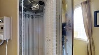 This single-room tanning space is purpose-built to accommodate a professional stand-up tanning unit. The centerpiece of the room is a vertical tanning booth enclosed by a structured grid panel lined with rows of tanning lamps, designed to provide even, all-around UV exposure. An integrated ceiling-mounted fan sits directly above the unit, ensuring proper ventilation and user comfort during sessions. The booth features a hinged or sliding access panel, allowing easy entry and exit. The surrounding walls have a warm, light yellow finish, contributing to a clean and functional atmosphere. A frosted window positioned nearby allows natural light to filter into the room without compromising privacy. The layout is compact and efficient, with the tanning unit taking up the primary floor space, leaving a small area for users to prepare before and after sessions. The room appears designed strictly for tanning purposes, with its equipment and layout optimized for safe, effective use. Overall, the space is utilitarian in design, prioritizing functionality and user accessibility within a well-maintained, single-purpose environment.