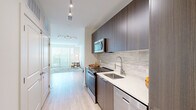 This single-floor space showcases a clean, contemporary design throughout. The galley-style kitchen is well-appointed with dark wood flat-front cabinetry extending to ceiling height, stainless steel appliances including a refrigerator with ice maker, electric range, built-in microwave, and dishwasher. Light quartz countertops contrast the cabinetry, while a subtle tile backsplash adds texture. Track lighting runs the length of the kitchen corridor, illuminating the light wood-style flooring that flows throughout the unit.

The living area is bright and open, anchored by a large sliding glass door that brings in generous natural light and outdoor views. White walls and clean lines keep the aesthetic minimal and versatile. A second flexible room features a frosted glass interior door and a large lean mirror, offering adaptable use as a bedroom or den.

The bedroom includes a sliding double-panel closet, providing practical storage within a calm, neutral setting. The bathroom features large-format beige floor tiles, a soaking tub with a marble-style surround, a floating vanity with dark wood cabinetry, and brushed nickel fixtures, completing the space with a polished, cohesive finish.