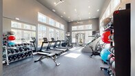 Located at 555 Linger Longer Dr on Johns Island, SC, this fitness center offers a thoughtfully designed workout environment within a single, open-plan space. The room is defined by striking vaulted ceilings that create an airy, expansive atmosphere, complemented by dual ceiling fans and recessed lighting throughout. Large floor-to-ceiling windows line multiple walls, flooding the interior with abundant natural light and providing views of the surrounding landscape — a feature that makes the space feel even more open and inviting.

The flooring consists of durable rubber gym matting in a speckled grey finish, providing both safety and functionality underfoot. Cardio equipment is prominently featured, with a row of BodyCraft treadmills, elliptical trainers, and spin bikes positioned to take advantage of the natural light and outdoor views. A stair-climber and cable/pulley strength machine add further variety to the workout options available.

The free weights section includes a full dumbbell rack ranging across multiple weight increments, accompanied by a flat weight bench and a full-length wall mirror that enhances the sense of space. A well-stocked ball rack holds medicine balls, stability balls, and slam balls in various sizes. Wall-mounted flat-screen televisions provide entertainment, while the grey-toned walls and clean white trim give the space a modern, polished aesthetic throughout.