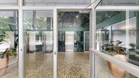 This elegant building lobby presents a striking mid-century modern design defined by an exceptional combination of premium materials and architectural precision. The space is enveloped by dramatic floor-to-ceiling glass walls that flood the interior with natural light and offer unobstructed views of lush, mature trees beyond. Rich green marble cladding lines the interior walls from floor to double-height ceiling, creating a bold and luxurious focal point that anchors the entire space. The polished terrazzo floors, featuring a multi-toned aggregate pattern, extend seamlessly throughout the hallways and lobby area, reinforcing the refined character of the design. A bank of stainless steel elevators is neatly integrated into the marble feature wall, providing clean, functional access. The lobby is furnished with classic Barcelona-style tufted leather chairs arranged around a low coffee table atop a patterned area rug, complemented by a sculptural sputnik chandelier that adds a distinguished decorative accent overhead. Tall tropical potted plants punctuate the space, softening the hard surfaces and introducing an organic warmth. Connecting hallways extend from the main lobby, maintaining the same high-quality material palette. The overall ambiance is polished, professional, and timelessly elegant.