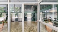 This elegant building lobby presents a striking mid-century modern design defined by an exceptional combination of premium materials and architectural precision. The space is enveloped by dramatic floor-to-ceiling glass walls that flood the interior with natural light and offer unobstructed views of lush, mature trees beyond. Rich green marble cladding lines the interior walls from floor to double-height ceiling, creating a bold and luxurious focal point that anchors the entire space. The polished terrazzo floors, featuring a multi-toned aggregate pattern, extend seamlessly throughout the hallways and lobby area, reinforcing the refined character of the design. A bank of stainless steel elevators is neatly integrated into the marble feature wall, providing clean, functional access. The lobby is furnished with classic Barcelona-style tufted leather chairs arranged around a low coffee table atop a patterned area rug, complemented by a sculptural sputnik chandelier that adds a distinguished decorative accent overhead. Tall tropical potted plants punctuate the space, softening the hard surfaces and introducing an organic warmth. Connecting hallways extend from the main lobby, maintaining the same high-quality material palette. The overall ambiance is polished, professional, and timelessly elegant.