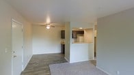 This single-floor residential space features a clean, open-concept design that seamlessly connects the living area to the kitchen. The living room is finished with warm, neutral-toned carpet and transitions into durable hardwood-style flooring, offering a comfortable and functional layout. A ceiling fan with a light fixture serves as a central element overhead, providing both airflow and ambient lighting to the space.

The kitchen is accessible via a half-wall breakfast bar with a light-toned countertop, creating an open pass-through that enhances the sense of connectivity between rooms. Dark espresso-finished cabinetry lines the kitchen walls, providing ample storage and a contrast to the white appliances and light-colored countertops. Recessed lighting illuminates the kitchen area efficiently.

Throughout the space, neutral beige and white walls contribute to a bright, airy atmosphere. White baseboards and clean trim details are consistent across all areas, lending a polished, well-maintained appearance. The overall layout is practical and straightforward, making efficient use of the available floor plan. The open sightlines between the living and kitchen areas make the space feel larger and more inviting, ideal for everyday living.