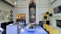 This single-story space in Dallas, Texas makes a striking first impression through its grand lobby design. The room is defined by dramatically soaring ceilings that draw the eye upward to a statement chandelier with ornate metalwork and exposed bulbs, suspended above a central arched architectural detail that adds depth and visual interest to the space.

The main seating area is anchored by a mirrored coffee table resting on a plush blue area rug, flanked by tufted gray sofas adorned with decorative throw pillows, creating an inviting and polished atmosphere. Opposite the main seating arrangement, a pair of charcoal accent chairs and a bold yellow tufted ottoman introduce contrast and vibrancy to the color palette.

A wide central corridor leads toward a glass-paned entryway, offering a sense of openness and connectivity throughout the space. Decorative wall art, topiary greenery, and carefully curated accessories contribute to the refined aesthetic. Along one wall, a textured partition backdrop adds dimension behind a secondary seating or display area. The overall ambiance is contemporary and upscale, blending elegant architectural elements with modern furnishings.