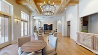 This thoughtfully designed single-floor space exudes a warm yet sophisticated ambiance, seamlessly combining transitional and traditional architectural elements throughout. Wide-plank hardwood floors flow continuously across the entire layout, anchoring each distinct area with a sense of cohesion and warmth.

The expansive main gathering area commands attention with its striking coffered ceiling accented by exposed natural wood beams. A large wrought-iron candle-style chandelier serves as a dramatic focal point overhead, while an abundance of tall windows and French doors floods the space with generous natural light and provides views to the surrounding exterior. Multiple conversational seating arrangements, featuring neutral-toned upholstered chairs and sofas, are thoughtfully positioned throughout to encourage intimate gatherings.

A dramatic library lounge anchors one end of the space, distinguished by floor-to-ceiling dark-painted built-in shelving, a plush navy velvet sofa, and curated artwork — creating a moody, intimate contrast to the airy brightness of the main areas. Adjacent sitting nooks offer quieter retreats, one featuring a rustic wood art piece and layered neutral furnishings.

A secondary lounge area benefits from soaring vaulted ceilings with exposed wood beams, wall-mounted sconces, and direct access to an outdoor courtyard through multiple French doors, enhancing the sense of openness and connectivity to the outdoors throughout this elegantly appointed space.