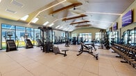 Located at 112 Bilby Road in Hackettstown, NJ, this generously sized fitness center offers a thoughtfully designed, single-floor layout that accommodates a full range of workout activities. The main workout hall is defined by a dramatic vaulted ceiling adorned with exposed natural wood beams, wood-paneled cladding, and integrated linear LED lighting that creates a bright, energizing atmosphere. Skylights and expansive floor-to-ceiling windows along multiple walls flood the space with natural light while providing open views of the surrounding outdoor amenities.

The main floor is finished with durable rubber sport tile flooring throughout and is equipped with an extensive array of strength training stations, including cable machines, multi-functional racks, free weight benches, and a full dumbbell rack displayed along a mirrored wall. Cardio equipment — including treadmills, ellipticals, and stationary bikes — is arranged to take advantage of the outdoor views. A dedicated functional training area features a heavy bag, TRX suspension system, rowing machine, and stability equipment, complemented by a wall-mounted television. A separate glass-enclosed studio area provides additional space for group or individual workouts. The overall ambiance is modern, clean, and professionally appointed, making this facility a comprehensive destination for fitness and wellness.