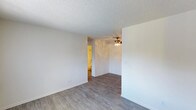 This single-floor space presents a clean and straightforward interior design with a neutral color palette throughout. The main living area features smooth, light-toned wood-look laminate flooring that adds warmth and continuity to the space. Bright white walls and crisp white baseboards contribute to an open, airy feel, while the textured popcorn ceiling adds a classic touch overhead. A ceiling fan with a light fixture is centrally mounted, providing both airflow and ambient lighting to the room. An open doorway leads to an adjoining room, which appears to be a bathroom or utility area based on its warm-toned lighting and tiled or vinyl flooring visible through the passage. The layout is compact yet functional, with a clear, unobstructed flow between spaces. The overall ambiance is clean, minimal, and move-in ready, making it a versatile space well-suited for a variety of uses. The simplicity of the finishes and the neutral tones throughout create a blank canvas that can easily be personalized to suit individual preferences.