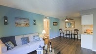 This single-floor space presents a cohesive coastal aesthetic, anchored by soft blue-painted walls that create a relaxed and airy atmosphere. The open-concept layout connects the living and dining areas seamlessly, promoting an easy flow between the two spaces. Rich, dark hardwood-style flooring runs throughout, providing a warm contrast to the cool wall tones and tying the rooms together visually.

The living area is comfortably sized, accommodating a plush white sofa accented with blue patterned throw pillows, complementing the nautical theme. A glass-top coffee table and decorative lantern add layered visual interest to the space. Beach-themed artwork adorns the walls, reinforcing the coastal character of the interior.

Adjacent to the living area, the dining space features a bistro-style table with curved chairs positioned beneath a ceiling fan with a light fixture, which provides both ventilation and ambient lighting. A decorative mirror nearby enhances the sense of space.

A doorway with louvered bifold doors leads to an additional room, likely offering storage or utility functions. The textured ceiling is consistent throughout, and HVAC vents are integrated discreetly into the ceiling for climate control.