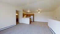 This single-floor residence offers a practical and comfortable open-concept layout that seamlessly connects the living area to the kitchen. The living room is generously sized, finished with soft carpeting underfoot and topped with vaulted ceilings that add a sense of airiness to the space. A ceiling fan keeps the area comfortable year-round, while flush-mounted lighting provides consistent illumination throughout.

The kitchen is well-appointed with warm, honey-toned wood cabinetry that provides ample storage space. A raised breakfast bar peninsula separates the kitchen from the living area, creating a natural divide while maintaining the open feel of the floor plan. The kitchen features tile flooring, standard appliances including a refrigerator and range, and an integrated exhaust hood above the cooking area.

Adjacent to the main living space, a hallway leads to a bathroom that features tile flooring and includes a vanity with cabinetry. A baseboard heater along the wall of the living room ensures efficient climate control throughout the colder months. The overall design is clean and functional, with a neutral color palette of whites and warm tones that creates a welcoming and versatile atmosphere suited to a variety of furnishing styles.