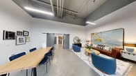 Located at 4015 133rd St SE in Mill Creek, WA, this single-floor loft-style unit offers a striking industrial-modern aesthetic defined by soaring ceilings with exposed ductwork, polished concrete floors, and an abundance of natural light streaming through large black-framed glass doors and transom windows. The open main area seamlessly integrates a dining space and living room, where a sliding barn door adds a functional yet stylish architectural detail. The living area features a cozy seating arrangement anchored by a textured area rug, complemented by warm and eclectic décor. The kitchen is thoughtfully designed with wood-toned cabinetry, open shelving, a tile backsplash, quartz countertops, and stainless steel appliances including a range, refrigerator, and range hood. The bathroom offers a bold, spa-like atmosphere with deep navy accent walls, a shower, and open wood shelving for storage. A dedicated laundry area houses a full-size front-loading washer and dryer. Linear pendant lighting and clean white walls throughout enhance the bright, contemporary feel of this well-appointed space.