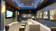 This single-level media lounge is designed to deliver an immersive entertainment experience in a stylish, theater-inspired setting. The room's most striking architectural feature is its tray ceiling, illuminated with vibrant blue ambient lighting that creates a dramatic cinematic atmosphere. Recessed can lighting complements the glow, ensuring the space is both functional and visually engaging.

The walls are painted in deep charcoal tones, reinforcing the moody, theater-like ambiance, while white baseboards provide a clean contrast. Abstract and contemporary framed artwork adorns the walls, adding a curated, lounge-like aesthetic. A wall-mounted flat-screen television anchors one end of the room, flanked by wall-mounted speakers for enhanced audio.

Seating is arranged to face the screen, consisting of plush striped sofas and loveseats paired with leather ottomans that offer flexible, casual comfort. A classic popcorn machine and a dedicated beverage cooler are conveniently positioned within the room, elevating the entertainment experience. The carpeted flooring in a neutral tone ties the space together, making this lounge a well-appointed destination for movie viewing and social gatherings.