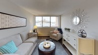This single-floor residence offers a thoughtfully arranged layout encompassing a living room, dining area, kitchen, bedroom, and bathroom. The living room is a welcoming space finished with dark hardwood-style flooring, neutral walls, and a large window fitted with blinds that allows natural light to fill the room. A baseboard heating unit runs along the exterior wall, and the space comfortably accommodates a full seating arrangement.

Adjacent to the living area is a dedicated dining nook, defined by the same flooring and neutral tones, providing an intimate setting for a four-person dining table beneath a flush-mount ceiling light. The galley-style kitchen is well-equipped, featuring white shaker cabinetry with brushed nickel hardware, gray countertops, a double stainless steel sink, and black appliances including a refrigerator and electric range, all set over wood-look flooring.

The bedroom offers plush carpeting, a wall-mounted air conditioning unit, a window with outdoor views, and a bi-fold closet with a built-in rod and shelving. The bathroom is finished with dark wood-look flooring, white cabinetry, a white countertop vanity, a large mirror, white subway tile accents, and a shower-tub combination.