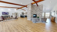 Located at 1001 Tramway Blvd NE in Albuquerque, New Mexico, The Reserve is a thoughtfully designed single-story community clubhouse offering a generous and versatile open-plan layout. The space is anchored by a dramatic floor-to-ceiling stacked stone fireplace that serves as a central architectural focal point, surrounded by comfortable seating areas ideal for gathering and relaxation. Rich wood beam accents traverse the vaulted ceilings throughout, lending warmth and character to the bright, airy interior.

Warm-toned hardwood-style flooring flows seamlessly across the entire space, unifying the distinct functional zones. The modern kitchen features gray cabinetry, stainless steel appliances, a large center island with bar seating, open shelving, and pendant track lighting — designed to support community events and casual socializing. Adjacent dining areas are bathed in natural light from large windows dressed with patterned curtains, offering views of the surrounding outdoor spaces.

A welcoming front reception area, branded with "The Reserve" signage, greets visitors with a sleek gray counter and clean white walls. Double doors lead to additional amenity spaces, including what appears to be a fitness or activity room. The overall ambiance is modern, warm, and inviting — reflecting a well-appointed community gathering destination.